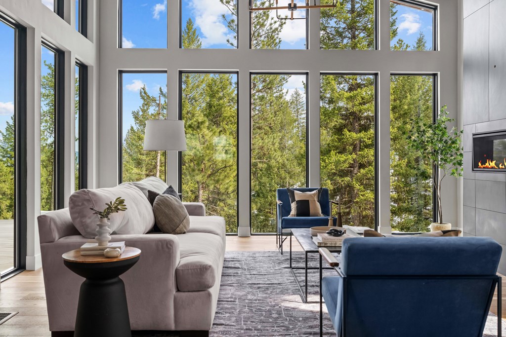 Bright, open living room with soaring windows, vaulted wood ceiling, and sleek black tile fireplace. Forest views fill the space, showcasing this mountain modern home’s signature design.