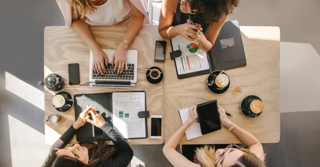 A group of women collaborate at a desk over coffee discussing real estate trends and marketing ideas for eXp Realty