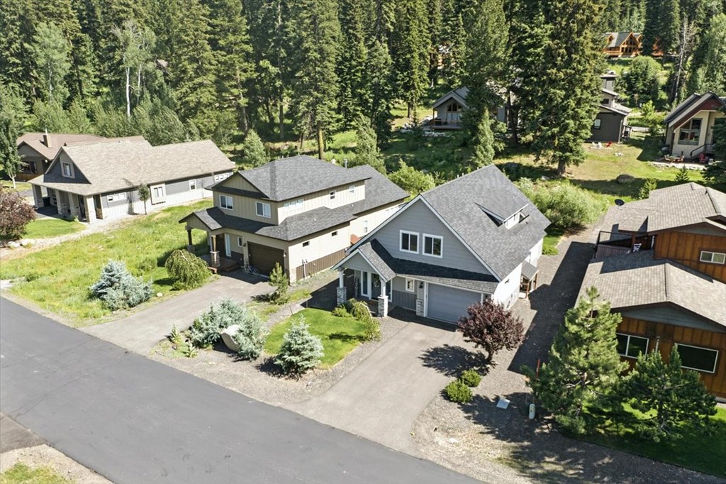 Aerial view of a McCall, Idaho home at 1484 Dragonfly Loop featuring modern gray siding, a two-car garage, and landscaped yard surrounded by mature pine trees and neighboring mountain-style homes. This McCall real estate property sits in a quiet wooded neighborhood close to downtown and outdoor recreation.
