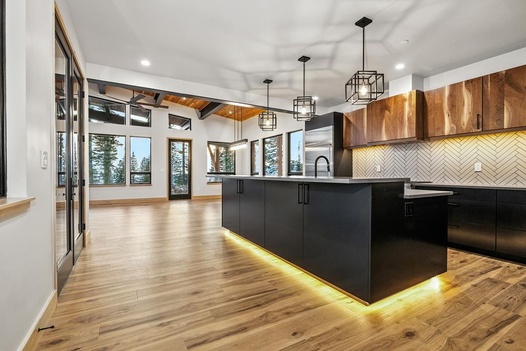 Open-concept mountain kitchen with large black island, wood floors, wall of windows, and vaulted timber ceiling.