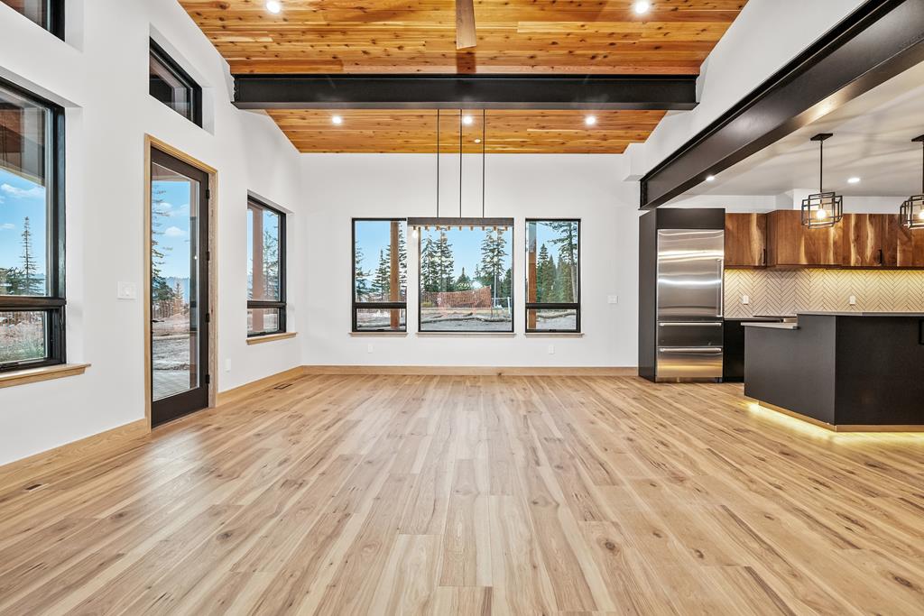 Bright great room with wide plank wood floors, black beams, and expansive windows looking out to the trees.
