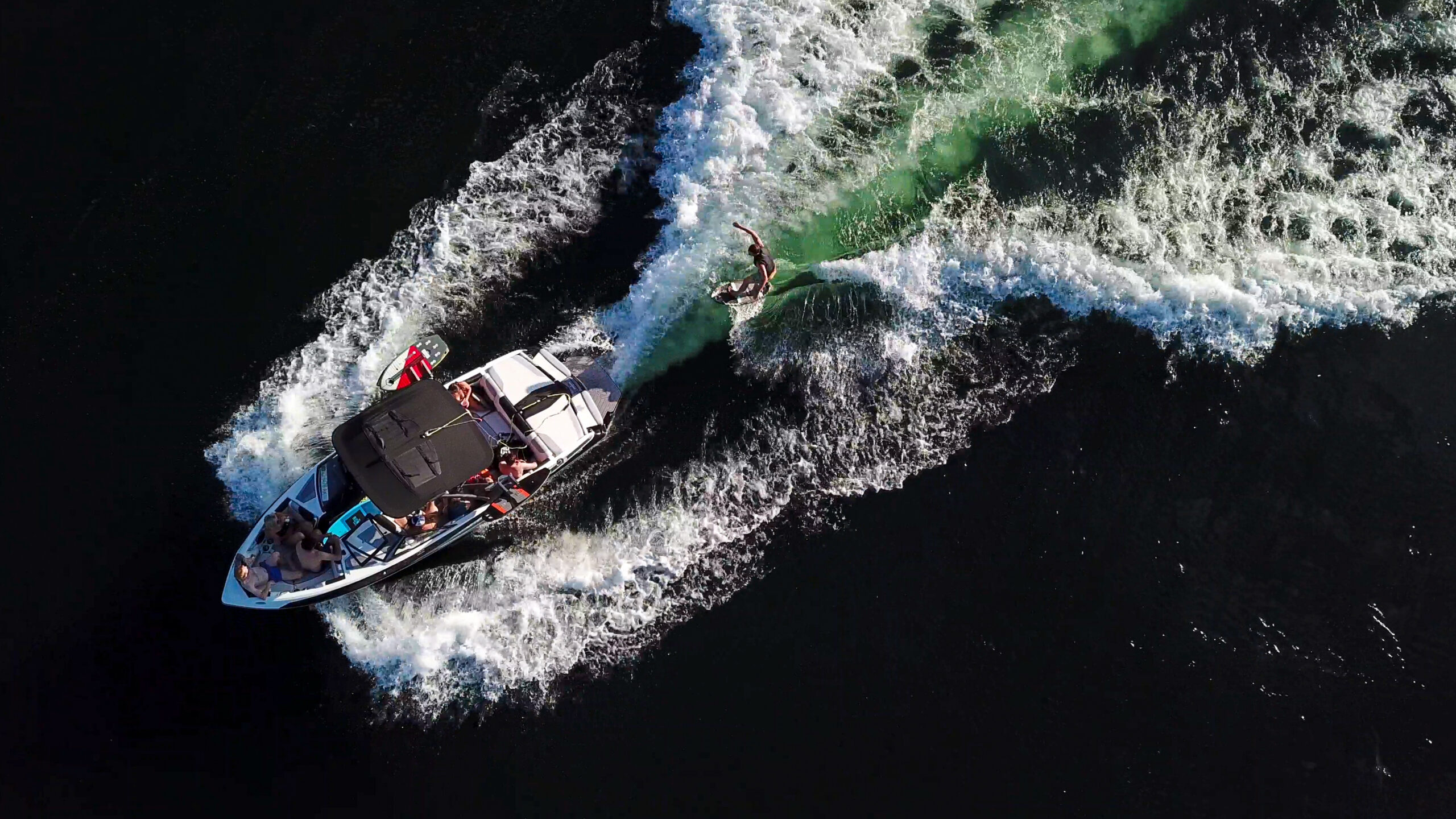 Aerial view of a wake surf boat pulling a surfer across dark lake water, leaving white waves behind.