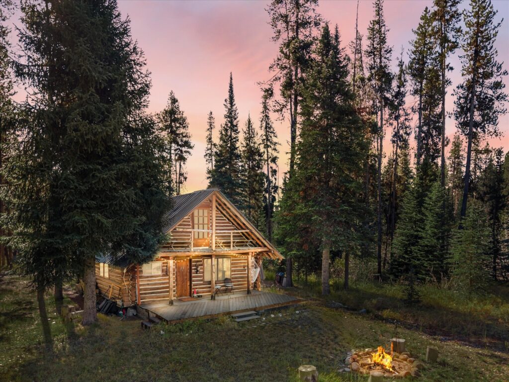 Log cabin at dusk under a pink sky, with a fire burning in the fire ring out front