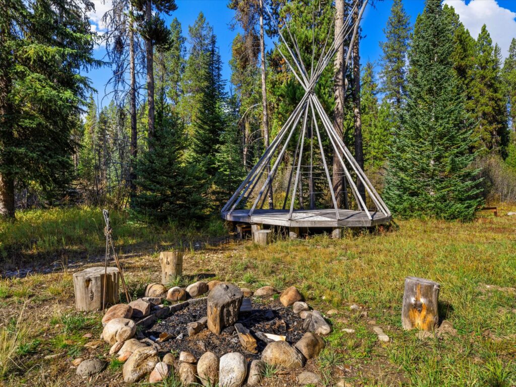 Stone fire pit with stump seats and a teepee frame on a wood platform in the clearing near the river