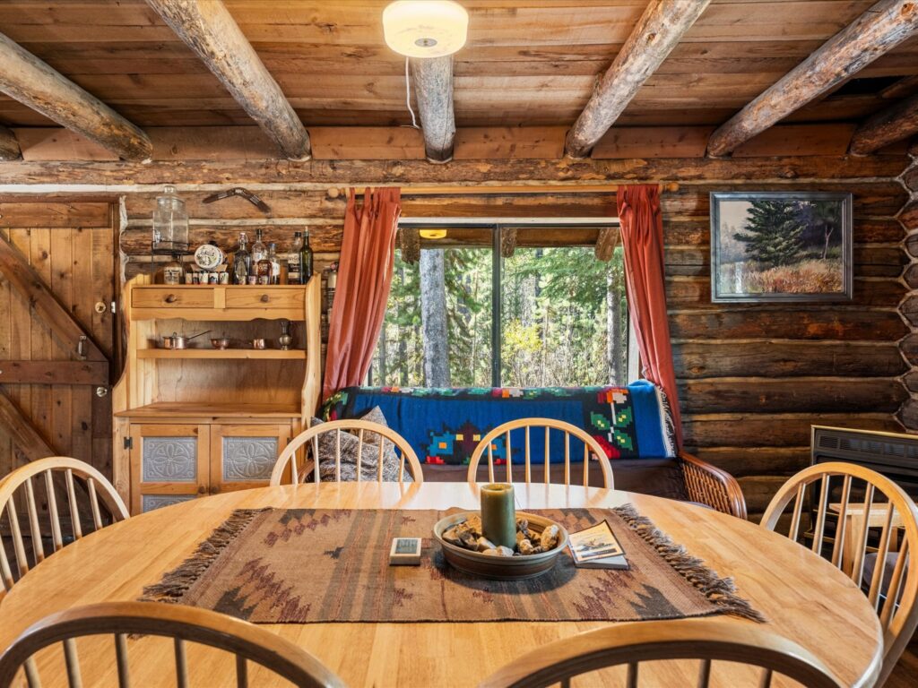 Round wooden dining table for six beneath exposed log beams, with a window onto the forest